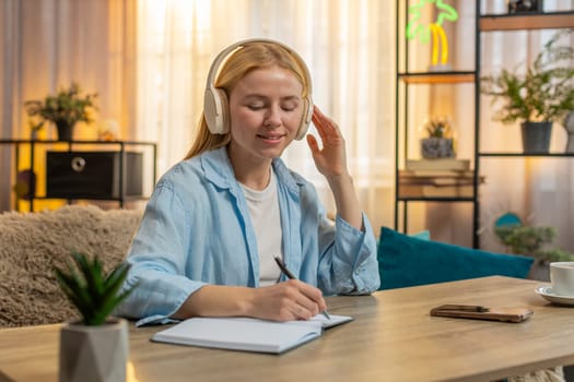 Young woman studies at home using headphones, listening an online course with bright curiosity. Caucasian girl at table records notes, combining comfort and skill improvement in sunset, motivation.