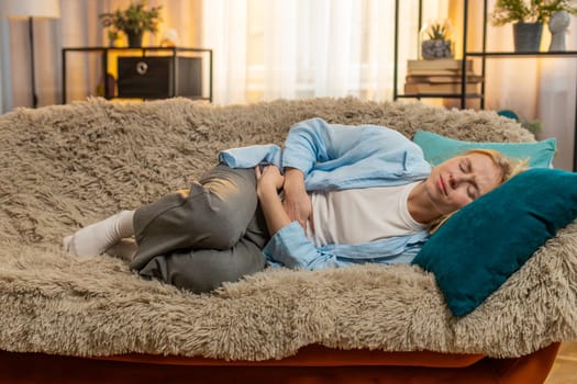 Young sick woman grabs abdomen at home, revealing cramps and grimaces from digestive turmoil. Caucasian girl on sofa lying in protective position, enduring painful tension and seeking calm relief.