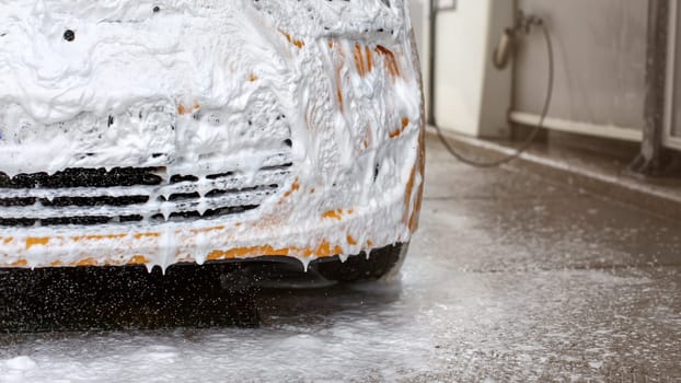 Front bumper of yellow car being washed in carwash, completely covered in white shampoo and foam, some dropping to the ground.