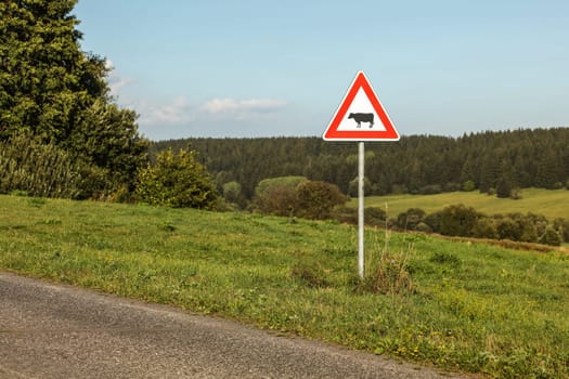 Caution cows crossing sign next to country road, forest in distance.