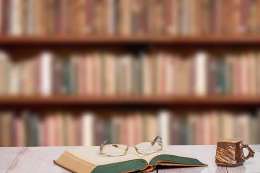 Books backgrounds. Closeup of an antique book with reading glasses on it and a brown clay mug on a light pink desk over a blurred bookshelf in library. Education concept. Teachers day.