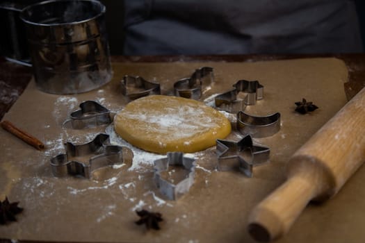 There is a dough on the parchment, around which there are cookie cutters of different shapes, all sprinkled with flour. High quality photo