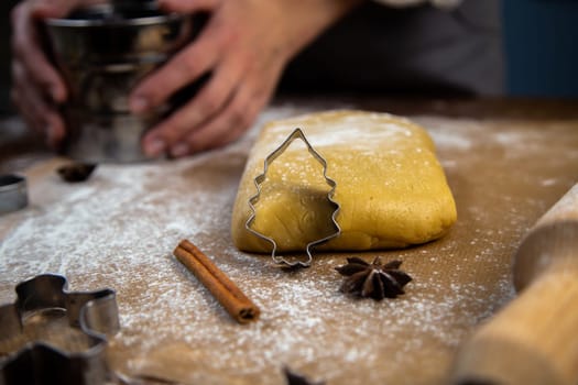 A cookie cutter in the form of a Christmas tree is leaning against the dough, a photo in a dark key, other molds are visible, a rolling pin, an iron sifter for flour, which is held in the hands of a pastry chef. High quality photo