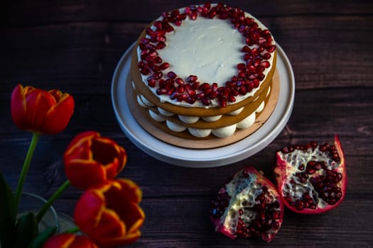 A cake of three layers consisting of cakes and white cream, decorated with pomegranate on top in a circle, stands on a dark background, next to tulips and pomegranates. High quality photo