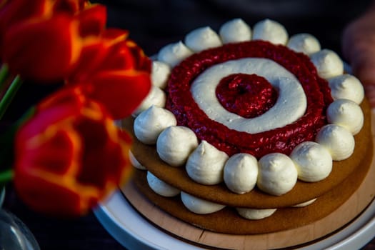 The baked cake cake lies on a wooden substrate and on a white stand, the cake is decorated with white cream and raspberry jam, in a circle, circles alternate, a bouquet of red tulips on a dark background is visible nearby. High quality photo