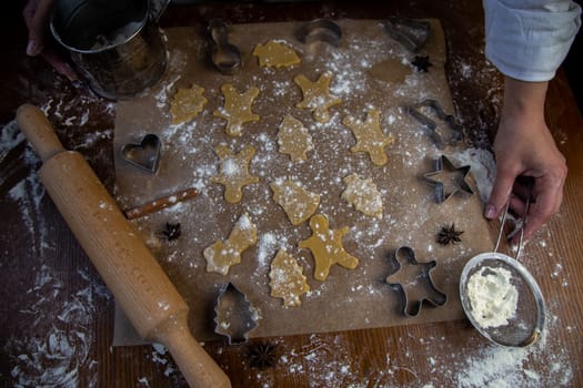There are ready-made cookies of various shapes on the baking parchment, a rolling pin, cookie cutters and a sieve with flour are lying nearby, flour is scattered everywhere, the hands of the pastry chef are visible. High quality photo