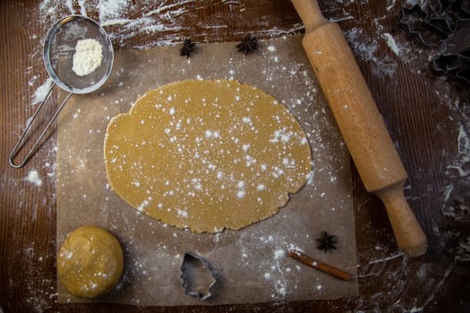 There is a rolled dough on the parchment, next to it is a lump of dough, a rolling pin, cookie cutters, cinnamon, everything is strewn with flour on a dark background. High quality photo
