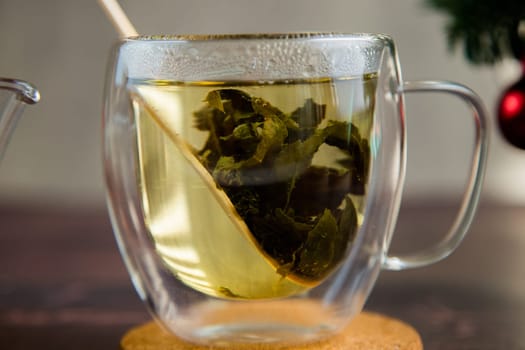 A transparent cup of tea stands on a wooden stand, green tea leaves and a wooden spoon float in the mug. High quality photo