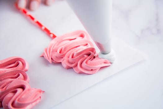 On light parchment there are pink meringue caps on sticks, the cook makes a pompom and a frill from a pastry bag of white meringue. Photography in a light key
