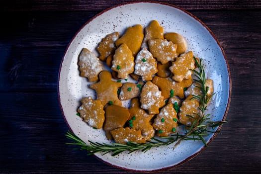 Cookies are on a plate of different shapes, in the foreground there is a cookie in the form of a Christmas tree, decorated with powdered sugar like snow and small green Christmas trees against the background of the rest of the cookies, a branch of rosemary is visible from the side. High quality photo