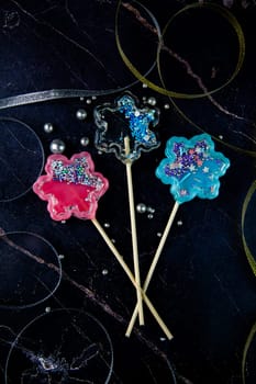Lollipops on a wooden stick in the form of a rattle in the form of transparent, pink and blue snowflakes lie on a dark marble background surrounded by gold and silver ribbons and silver balls. vertical photography, High quality photo