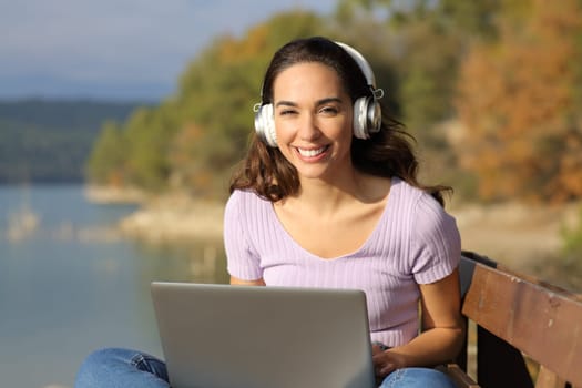 Front view portrait of a happy woman with laptop and headset looks at you in nature