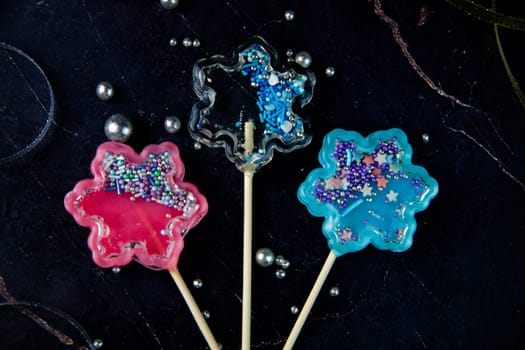 Lollipops on a wooden stick in the form of a rattle in the form of transparent, pink and blue snowflakes lie on a dark marble background surrounded by gold and silver ribbons and silver balls. High quality photo