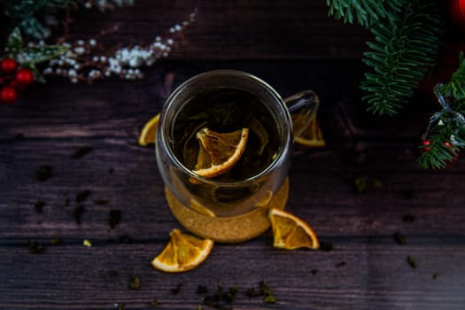 A transparent cup of tea stands on a dark table, on a wooden stand, dried orange slices float in the cup, citrus slices lie next to it, tea is scattered, a photo in a dark key. High quality photo