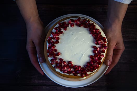 A cake of three layers, consisting of cakes and white cream, decorated with pomegranate on top, is held in the hands of a cook on a dark background, top view. High quality photo