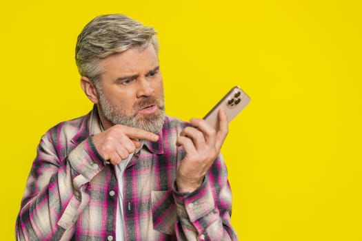 Mature man speaks on mobile phone with forceful tone, revealing anger agitation during tense talk quarrel. Middle-aged guy on yellow background shows argumentative stance, emphasizing heated dispute.