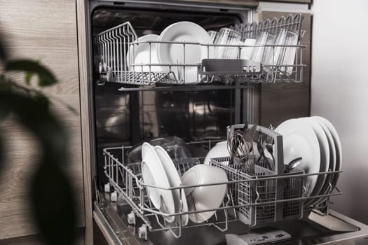 Front view of open automatic stainless steel built-in fully integrated top control dishwasher range machine with clean utensils, cutlery, glasses, dishes, plates inside in modern home kitchen