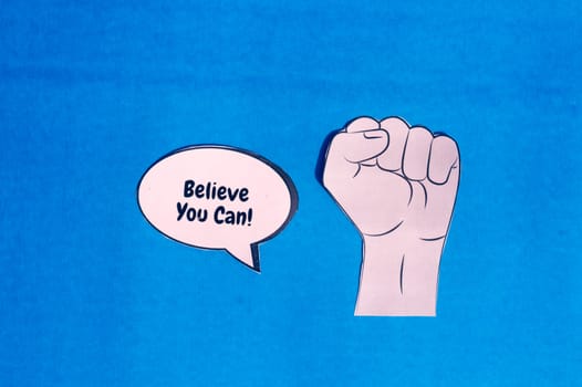 Firm human hand with motivation text on speech bubble isolated on blue background. Conceptual image shows that the first thing is to believe you can to achieve your goal.