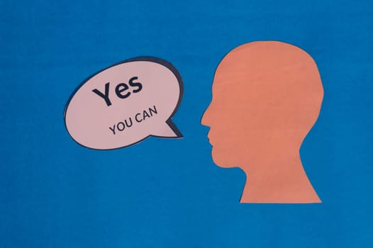 Paper human face with motivation text Yes You Can on speech bubble isolated on blue background. Conceptual image speaker acknowledges that you have the capacity, strength and determination to do.