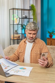 Caucasian mature man at home, using laptop and smartphone, reviewing financial reports. Middle-aged guy appears exhausted, rubbing eyes and diligently trying to find solutions for financial problems