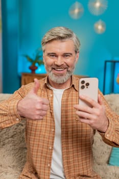 Mature man sitting on sofa at home, holding smartphone, giving thumbs up like gesture. Middle-aged guy shows approval on social media, smiling happily, enjoying positive content good game application