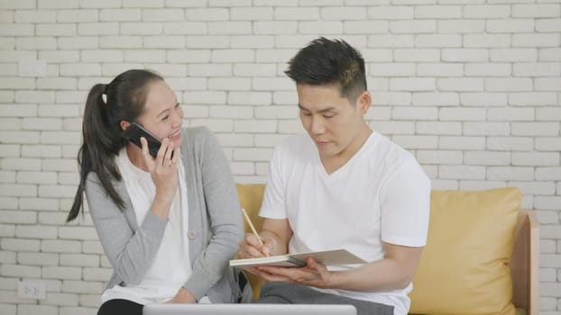 Happy Asian family couple husband and wife working confirming booking having talking call phone conversation and making writing notes of information together from home in living room. small business