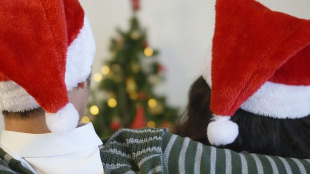 Happy Asian beautiful young family couple husband and wife sitting on sofa wearing a santa hat in front of fireplace at warm home Christmas winter hugging living room, back view