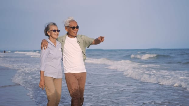 Happy Asian senior man and woman couple holding hands walking from the beach sunny with bright blue sky, Romantic elderly enjoy Travel summer vacation, plan life insurance at retirement couple concept