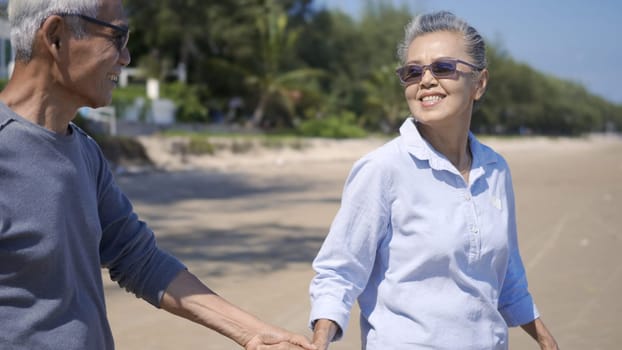 Happy Asian senior man and woman couple holding hands walking to the beach sunny with bright blue sky, Romantic elderly enjoy Travel summer vacation, plan life insurance at retirement couple concept