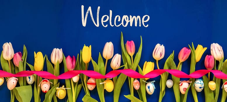 English Text Welcome. Banner Of White And Pink Tulip Spring Flowers With Ribbon And Easter Egg Decoration. Blue Wooden Background