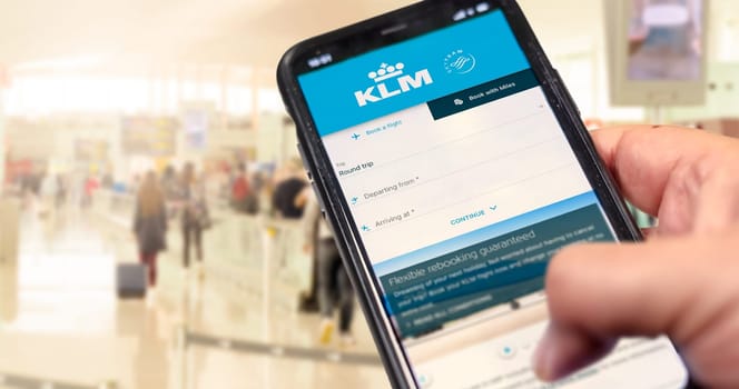 Amsterdam, NED, October 2021: Hand holding a phone with KLM Airlines flight booking application. Blurred airport with passengers in the background. Booking airline tickets for a round trip