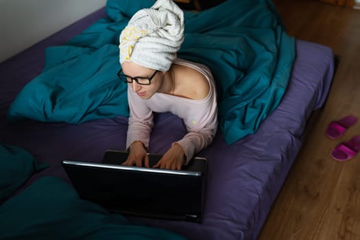 Home office, telecommuting, isolation, quarantine. A young girl working on a laptop, sitting on a bed at home in a towel after a shower