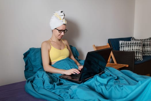 Home office, telecommuting, isolation, quarantine. A young girl working on a laptop, sitting on a bed at home in a towel after a shower