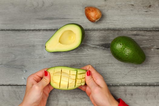 Tabletop view, young woman hands holding avocado with cuts, getting out the pulp.