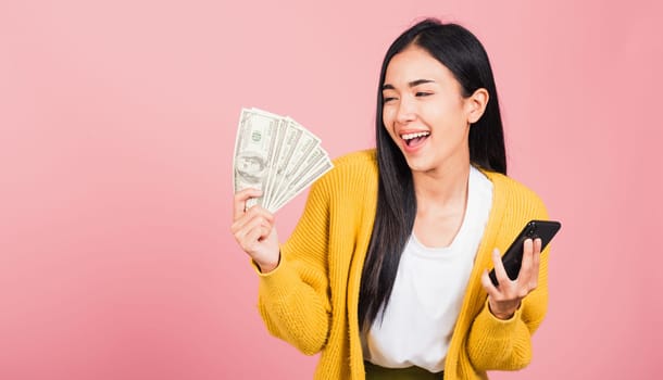 Portrait Asian happy beautiful young woman teen shopper smiling standing excited holding online smart mobile phone and dollar money banknotes on hand in summer, studio shot isolated on pink background