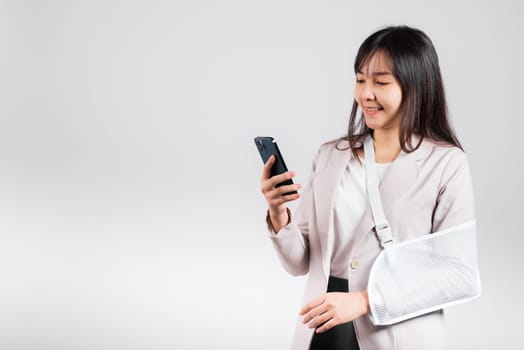 Woman confident smiling broken arm after accident and wear arm splint for treatment and hold smartphone, happy Asian female sling supported hand isolated on white background, social media, copy space