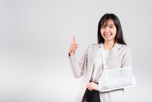 Arm broken. Business woman confident smile broken after accident wear arm splint for treatment show thumb up finger for good sign, happy Asian female sling support hand isolated on white background