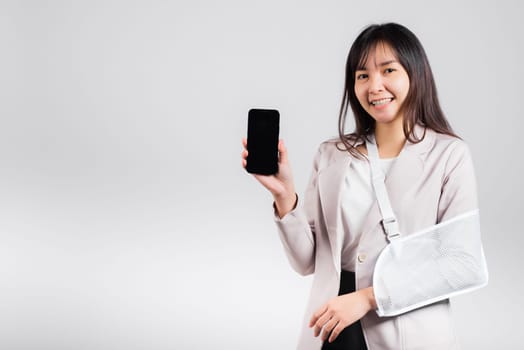 Woman confident smiling broken arm after accident and wear arm splint for treatment and hold smartphone, happy Asian female sling supported hand isolated on white background, social media, copy space