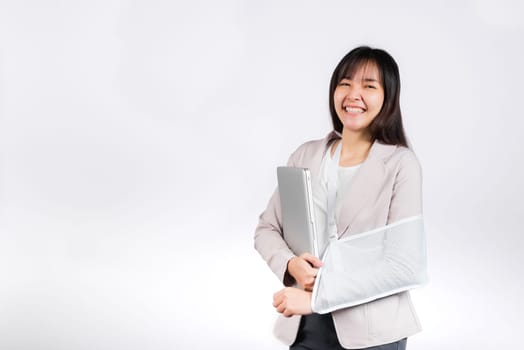 Asian business woman confident smiling broken arm after accident and wear arm splint for treatment but still have to work she hold laptop computer studio isolated on white background,copy space