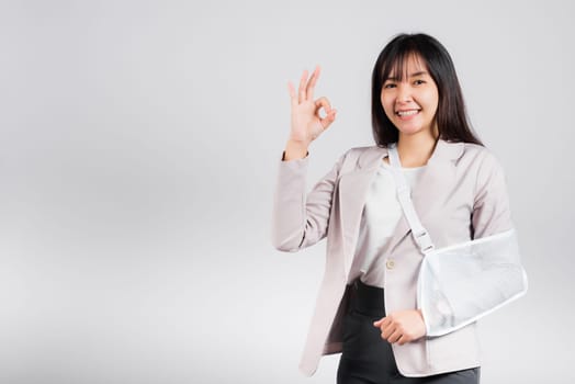 Arm broken. Business woman confident smile broken arm after accident wear arm splint for treatment show OK sign, happy Asian female sling support hand studio isolated on white background, copy space