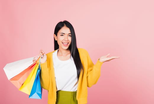 Portrait Asian happy beautiful young woman shopper smiling standing excited holding shopping bags multicolor in summer show something hand palm to copy space, studio shot isolated on pink background
