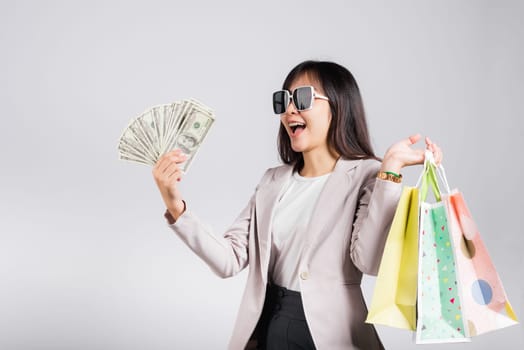 Woman with glasses confident shopper smile holding online shopping bags multicolor and dollar money banknotes on hand, excited happy Asian young female person studio shot isolated on white background