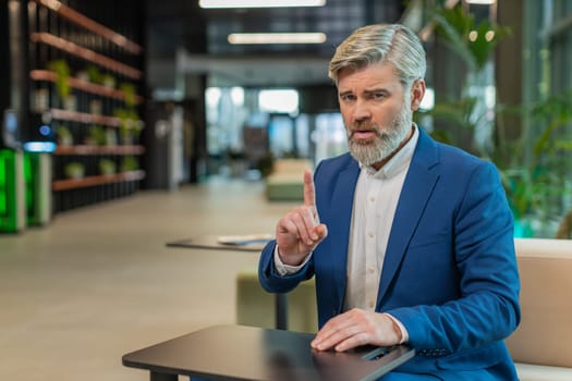 Portrait of mature businessman shakes finger and saying No be careful scolding and giving advice to avoid danger mistake disapproval sign sitting in modern office lobby. Serious guy in suit at hotel.