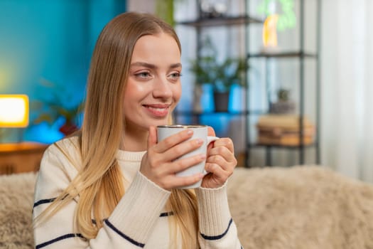 Caucasian woman girl sitting at home sofa holding hot tea cup relaxing enjoying quiet moment. Warm soothing drink brings calm cozy feeling indoors stress relief modern lifestyle content peaceful mood
