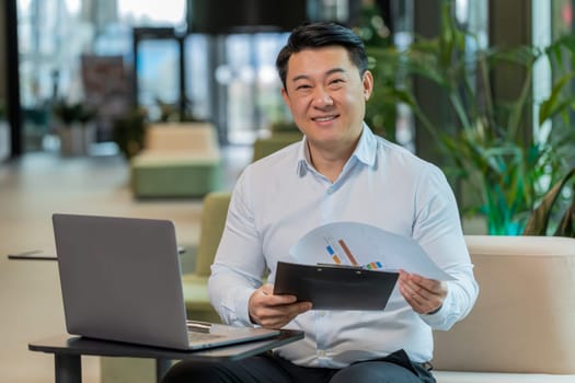 Chinese businessman working with documents, analyzing information, reading papers prepare financial report. Young male entrepreneur in formal using laptop analyzing graph documents in office lobby.