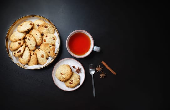 Homemade oatmeal cookies, tea cup, spoon, cinnamon stick and star anise on black table. Copy space for text. Flat lay.