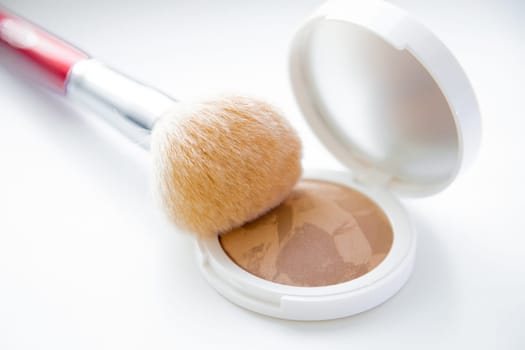 A white pillbox with powder and bronzer lies on a white background, on it lies a brush for blush on top. High quality photo