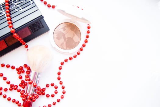 A white pillbox with powder and bronzer, a black package with eye shadow and a brush lie on a white background, they are entwined with red beads, with a place for text. High quality photo