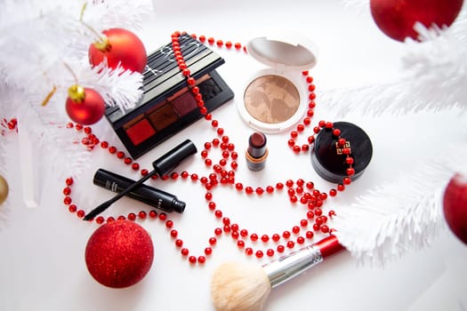 Powder and bronzer, eye shadow, lipstick, blush and a brush of different brands lie on a white background surrounded by white fluffy branches of a Christmas tree and red Christmas balls. High quality photo