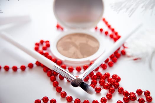 A white pillbox with powder and bronzer lies on a white background, thin makeup brushes and bright red beads lie in front. High quality photo